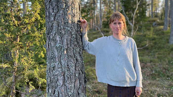 Blond kvinna med grå tröja och ena handen mot träd i skog. Foto